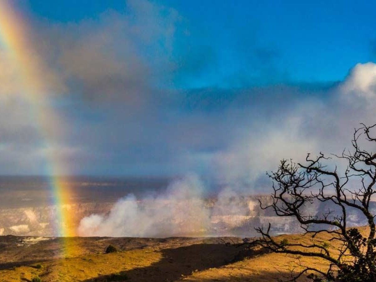 Rainbow over a volcanic landscape with smoke and a tree silhouette under a blue sky.