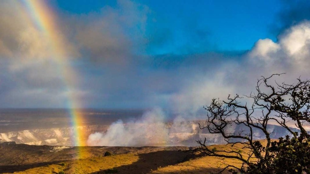 Rainbow over a volcanic landscape with smoke and a tree silhouette under a blue sky.