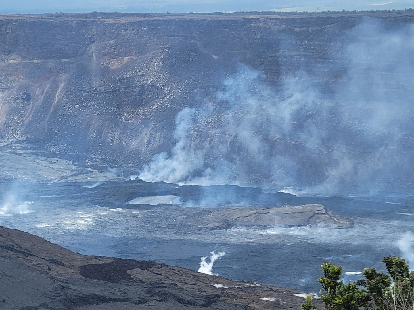 Volcanic crater with smoke rising, surrounded by rocky landscape and sparse vegetation.