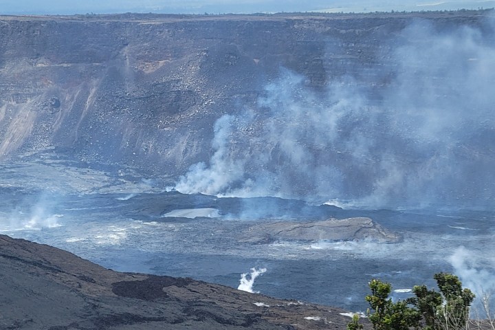 Volcanic crater with smoke rising, surrounded by rocky landscape and sparse vegetation.