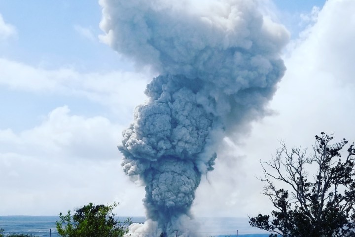 Eruption with large ash cloud rising into blue sky, surrounded by vegetation.