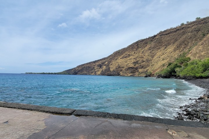 Rocky coastline and clear blue sea under a bright sky, with a steep hillside in the background.