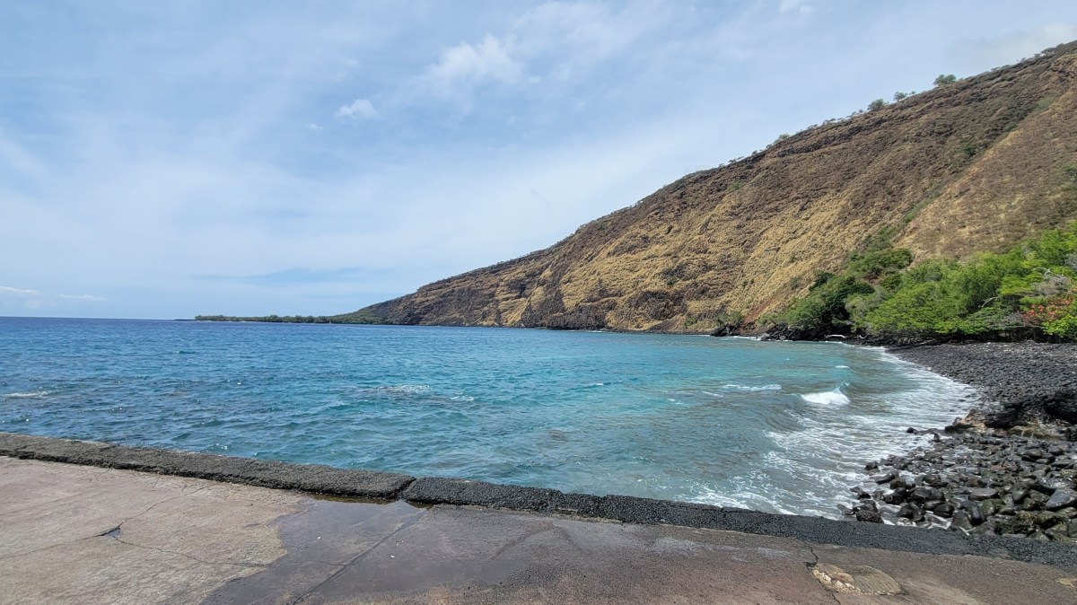 Rocky coastline and clear blue sea under a bright sky, with a steep hillside in the background.