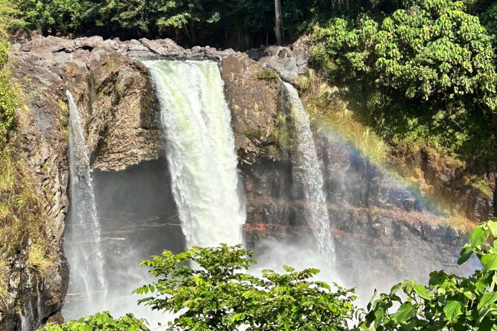 Waterfall cascading over rocks with a rainbow and lush green trees surrounding it.