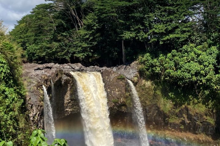Waterfall with rainbow and lush green trees under a cloudy sky.