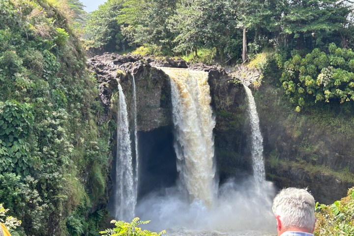 Waterfall cascading over a cliff surrounded by lush greenery, with people observing.