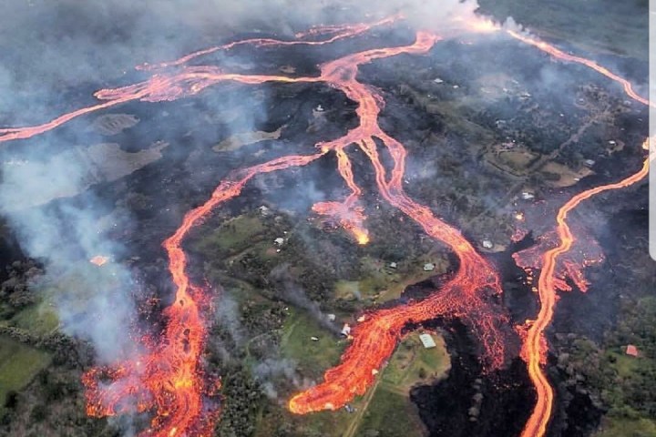 Aerial view of lava flowing from a volcano, surrounded by smoke and green landscape.