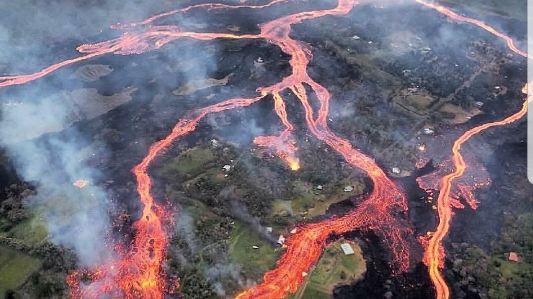 Aerial view of lava flowing from a volcano, surrounded by smoke and green landscape.